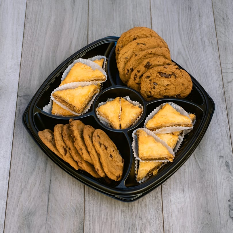 A black platter filled with triangular baklava pastries and chocolate chip cookies on a wooden surface.