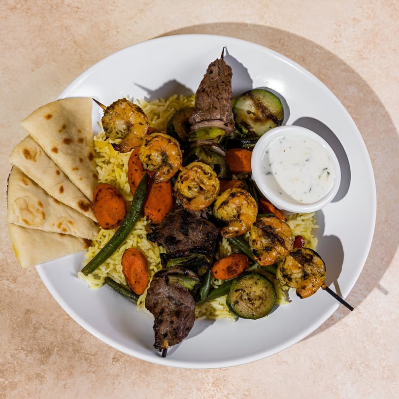 A Daphne's Greek Cafe plate with rice, grilled vegetables, shrimp and beef skewers, pita, and tzatziki.
