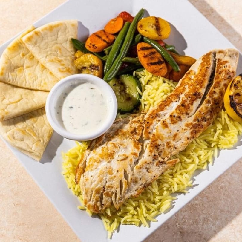 Grilled fish with yellow rice, grilled vegetables, pita bread, and a side of sauce on a white plate.